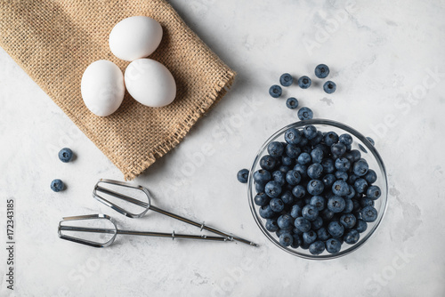 Top view photo of blueberries in a glass plate. Mixer fits on a white surface. Eggs on a brown bagging. Ingredients for healthy rustic breakfast.. Berries scattered of a surface.