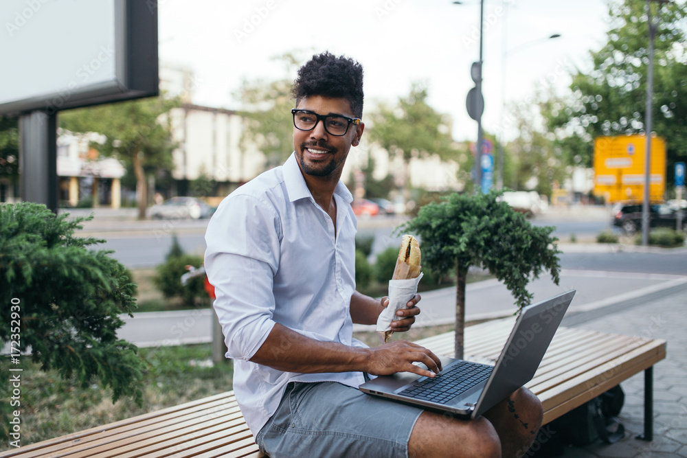 Young handsome Afro American man with glasses sitting on street with laptop and eating sandwich. Business concept. Young handsome Afro American man with glasses sitting on street with laptop and eating sandwich. Business concept.