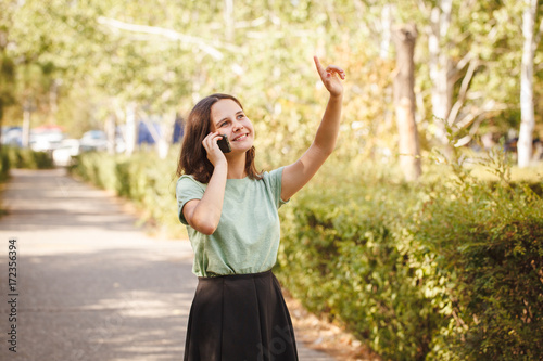 A young teenage girl outdoors talking on the phone, looking up and pointing a finger at something.