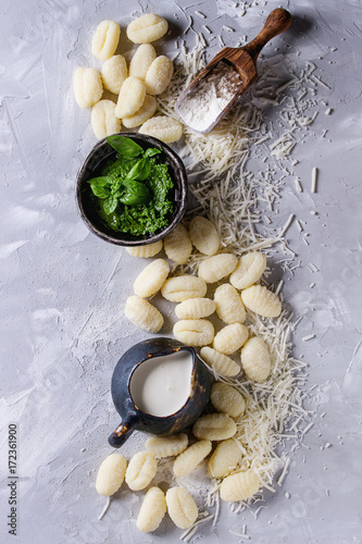 Raw uncooked potato gnocchi in black wooden plates with ingredients. Flour, grated parmesan cheese, basil pesto sauce, jug of cream over gray concrete background. Top view with space. Home cooking. photo