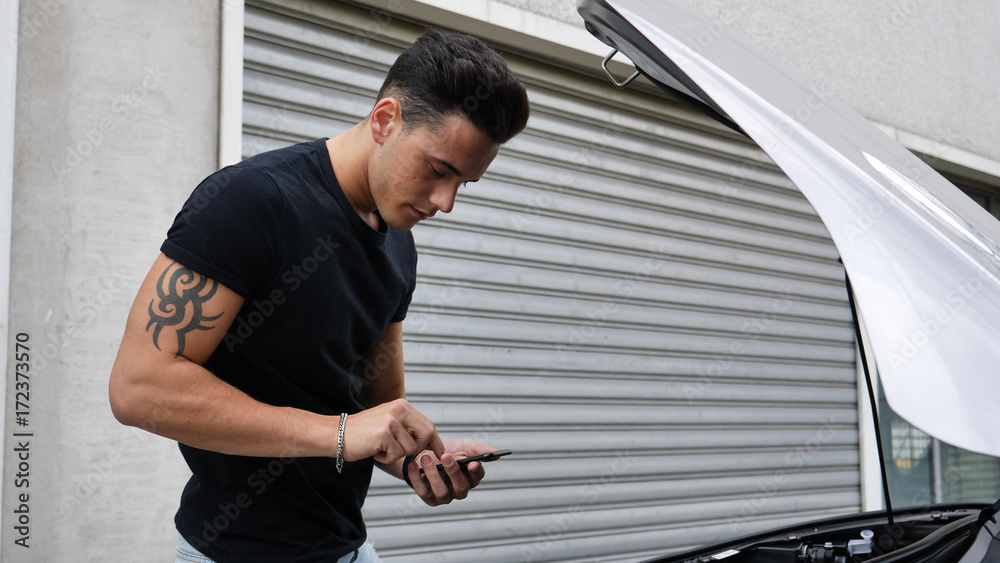 Fototapeta premium Young man trying to repair a car engine and seeking help with cell phone calling a mechanic