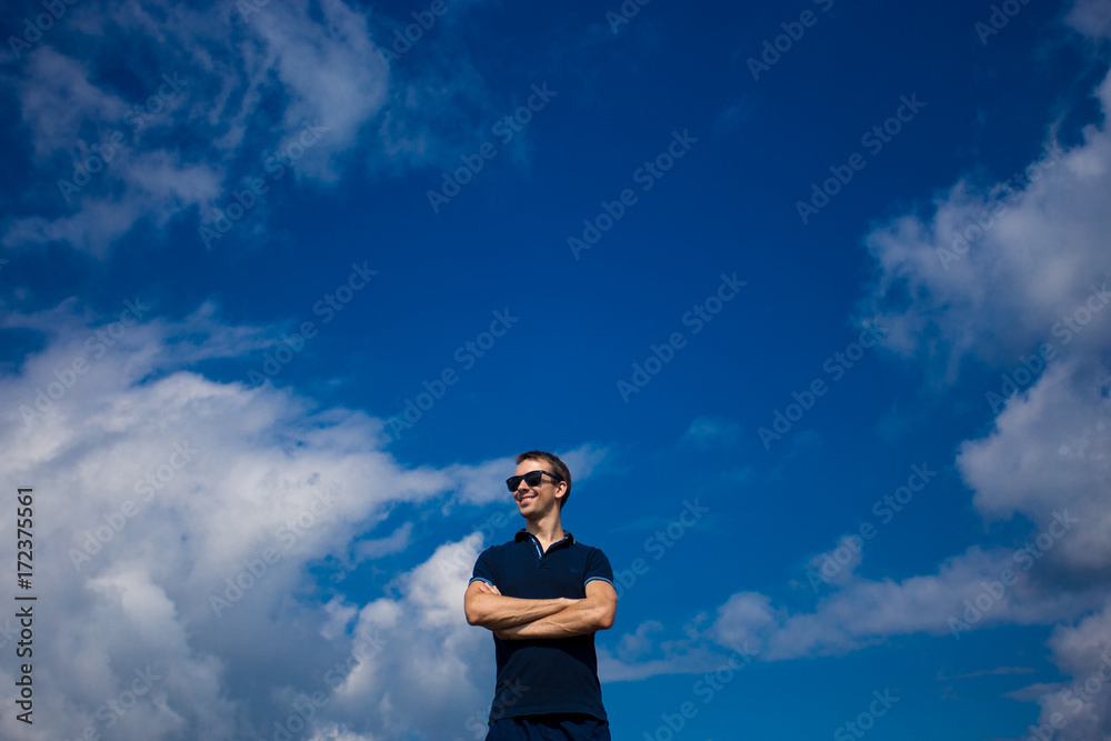 Young attractive man in sunglasses stands against the blue sky with arms folded Young attractive man in sunglasses stands against the blue sky with arms folded