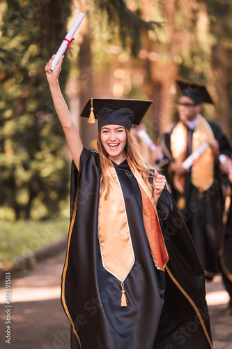 The young happy joyful graduate in mantle standing outdoor and keeping diploma in hand and smiling