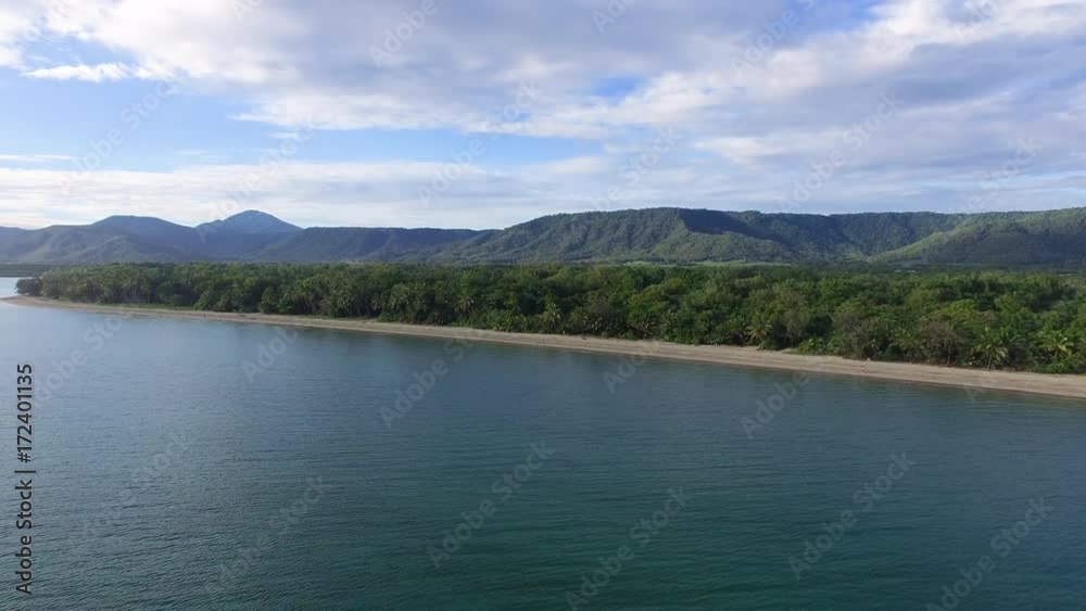 An aerial wide shot of a beautiful island. The island is covered with green tall trees. Mountain ranges is seen in the background under a cloudy weather. An aerial wide shot of a beautiful island. The island is covered with green tall trees. Mountain ranges is seen in the background under a cloudy weather.