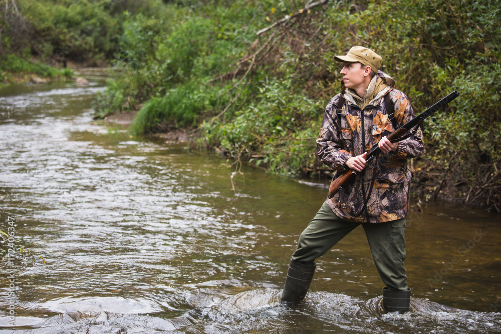 Hunter with a backpack and a hunting gun in the autumn forest. The man is on the hunt. The hunter crosses the forest river. Hunter with a backpack and a hunting gun in the autumn forest. The man is on the hunt. The hunter crosses the forest river.