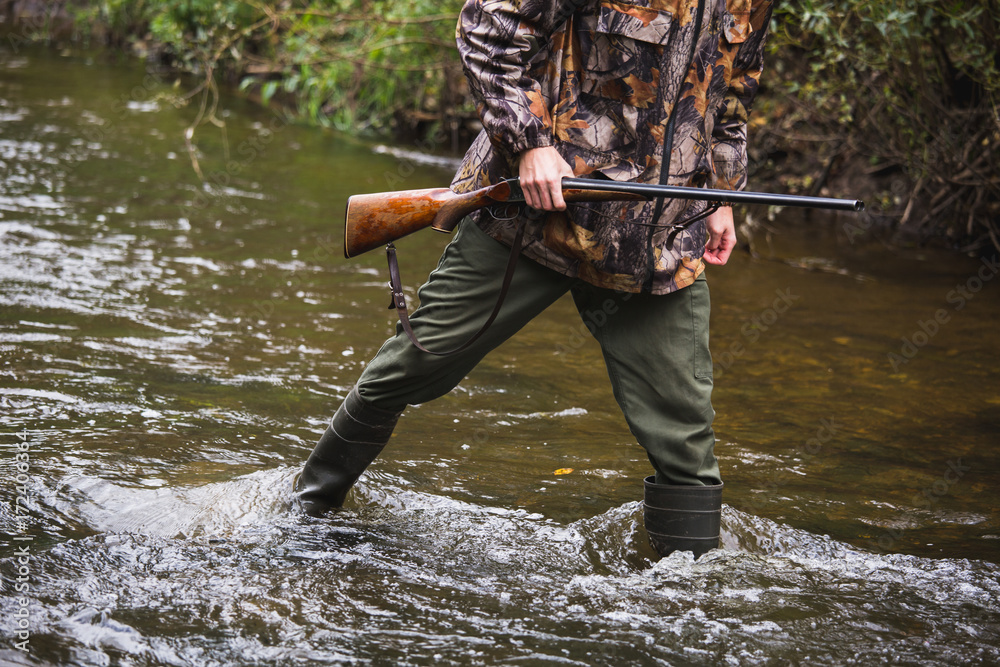 Hunter with a backpack and a hunting gun in the autumn forest. The man is on the hunt. The hunter crosses the forest river. Hunter with a backpack and a hunting gun in the autumn forest. The man is on the hunt. The hunter crosses the forest river.