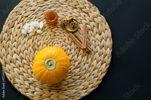 Small pumpkin with seeds  little glass can of honey  walnuts and cinnamon sticks on a circle mat napkin made of water hyacinth on a black background