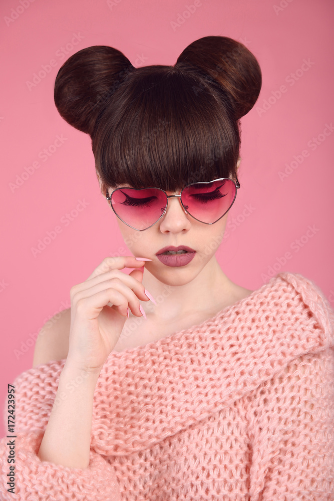 Fashion style teen girl model in knitted sweater. Brunette in heart sunglasses with matte lips and bun hairstyle posing over studio pink background. Fashion style teen girl model in knitted sweater. Brunette in heart sunglasses with matte lips and bun hairstyle posing over studio pink background.