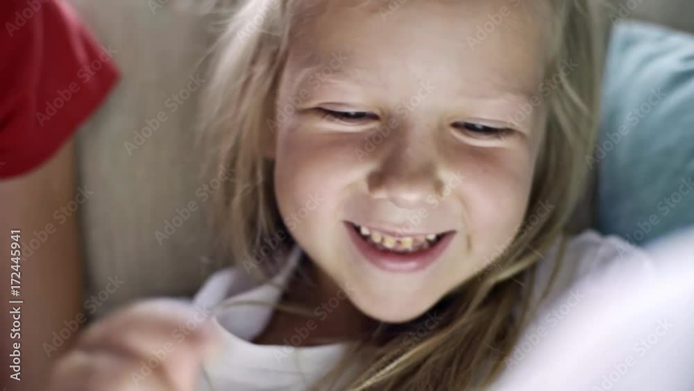 Close up of little girl with blond hair watching something and laughing while sitting on sofa with her sister Close up of little girl with blond hair watching something and laughing while sitting on sofa with her sister