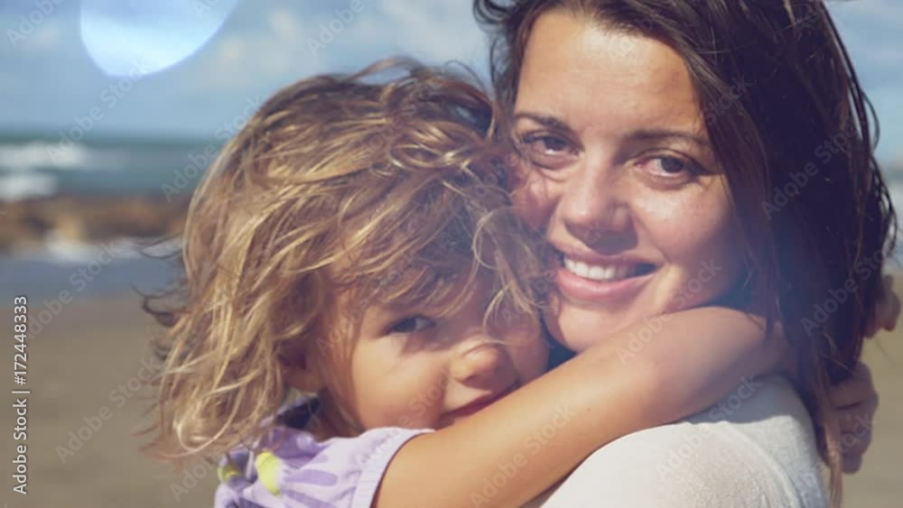 Beautiful woman holding in arms little blond daughter smiling in front of the ocean looking camera slow motion