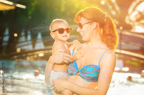 Mother holding a laughing child at tropical summer swimming pool background.