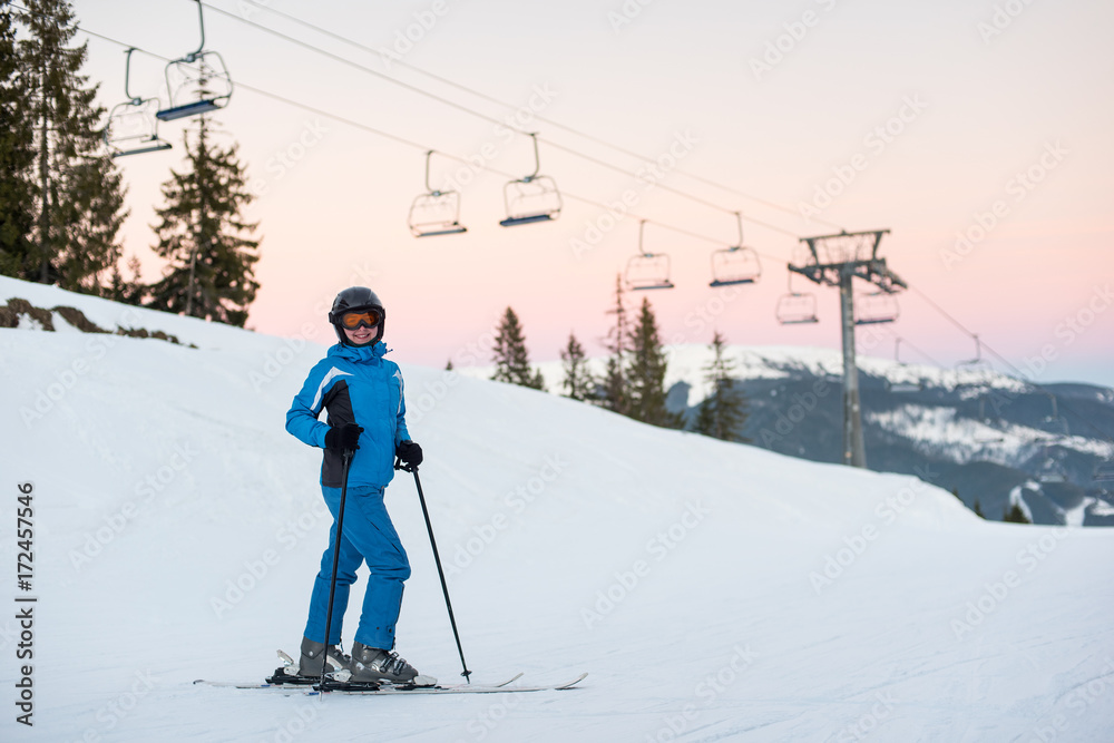 Happy skier female in blue jacket, ski pants and goggles on her head standing in the snowy mountains enjoying winter holidays against a ski-lift