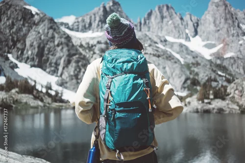 Obraz Kobieta wycieczkuje w śnieżnym halnym pustkowiu z plecakiem. Hipster wędrówki w pobliżu oszałamiającego jeziora lodowego w zabawnym kapeluszu pom