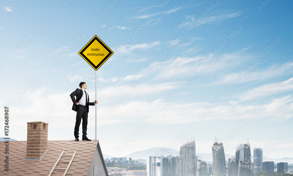 Young businessman on house brick roof holding yellow signboard and looking at city. Mixed media Young businessman on house brick roof holding yellow signboard and looking at city. Mixed media