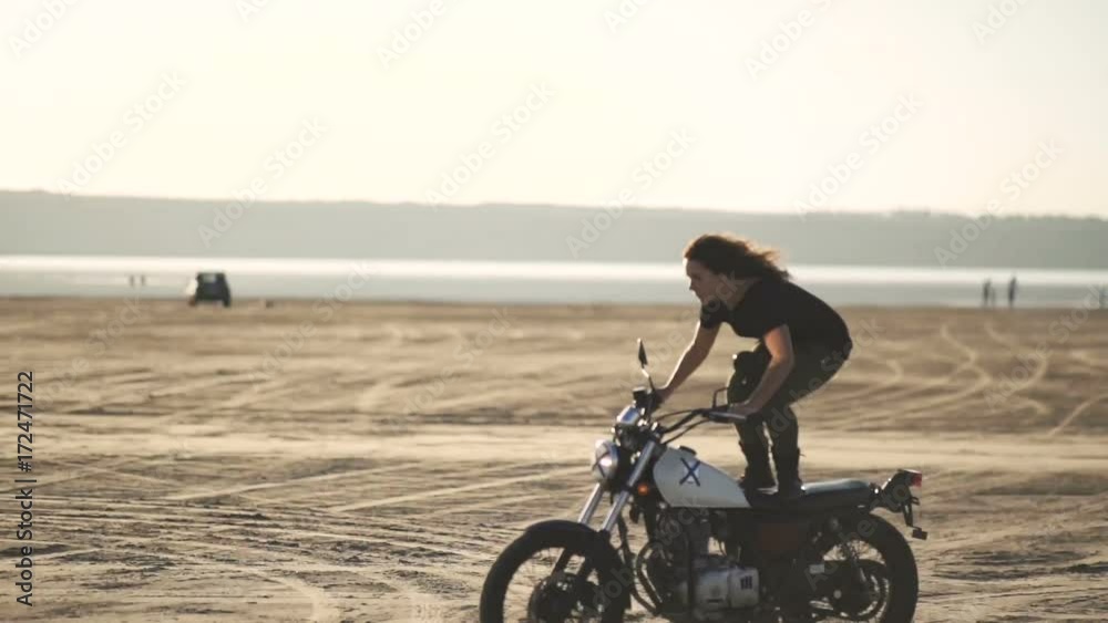  beautiful young woman riding a motorcycle rides on an old motorcycle and doing a trick. in the desert at sunset or sunrise. Female biker.