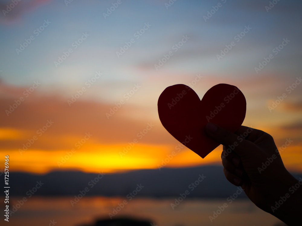 Silhouette hand is raising the red paper heart with blur sunlight during sunset, Valentines day.
