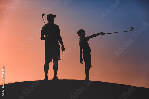 silhouettes of man with his son golfers standing with clubs on golf course at sunset