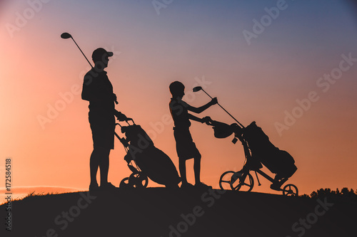 silhouettes of man with his son golfers walking with bags on golf course at sunset