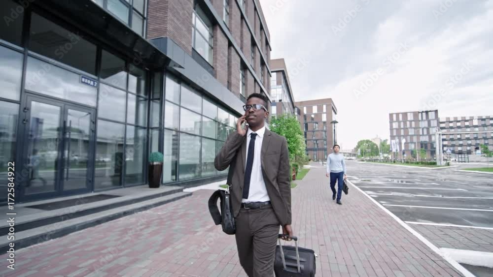 Slowmo of African businessman walking on street in downtown with suitcase and talking on mobile phone Slowmo of African businessman walking on street in downtown with suitcase and talking on mobile phone