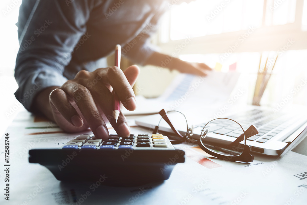 Close up of businessman or accountant hand holding pen working on calculator to calculate business data, accountancy document and laptop computer at office, business concept - obrazy, fototapety, plakaty 