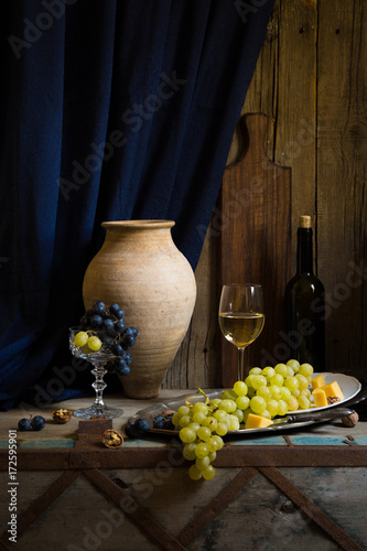 White wine in glass goblet green yellow grapes, bottle and pottery jar, silver tray cheese platter wooden board, blue curtain with drapes, vintage old tavern still life, family winery, eco farm