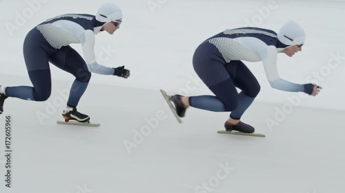 Tracking with slow motion of professional female speed skaters wearing spandex full-body covering suits sprinting along track in ice rink