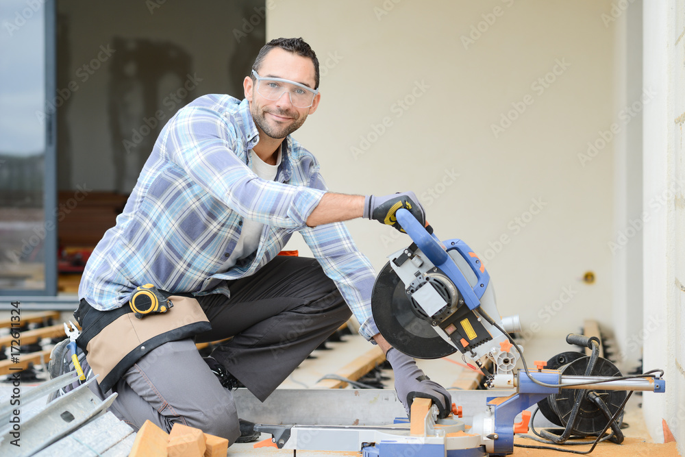 handsome young man carpenter using a circular saw while installing wood floor terrace outdoor in new house construction site
