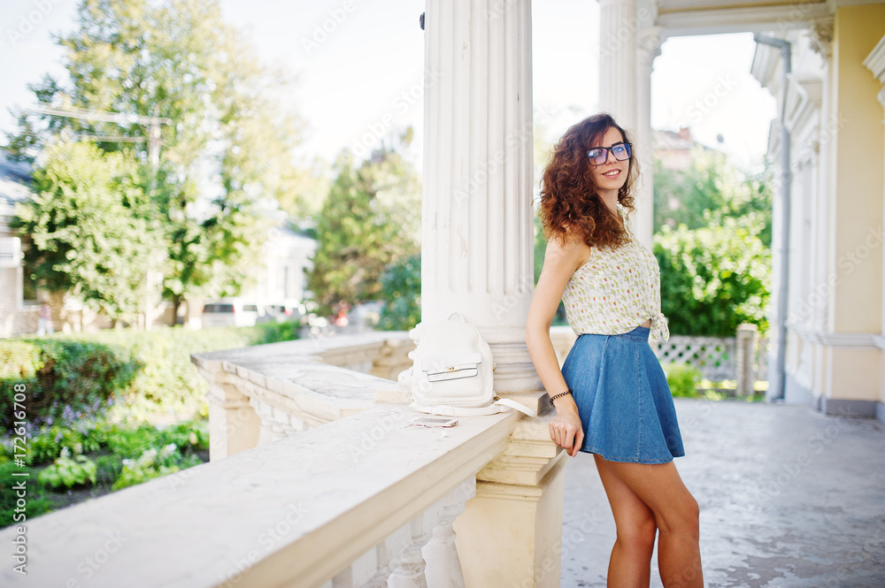 Curly stylish girl wear on blue jeans skirt, blouse and glasses posed near old vintage house. Curly stylish girl wear on blue jeans skirt, blouse and glasses posed near old vintage house.