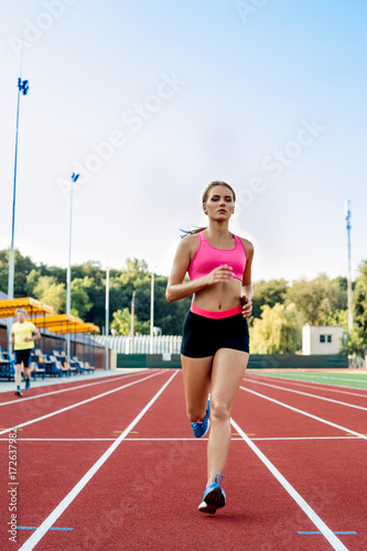 Sporty fitness woman jogging on red running track in stadium. Training summer outdoors on running track line with green trees on background. © nazarovsergey