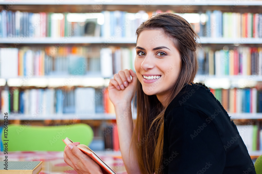 Young student girl in a library holding and reading a book sitting at table.