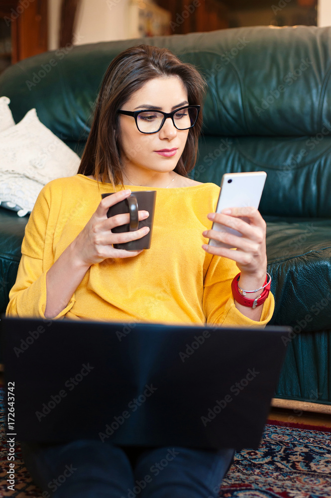 Woman sitting on the floor using a laptop and mobile phonem drinking coffee.