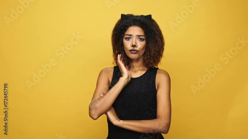 Young upset african woman in haloween costume with cat make-up and ears looking camera isolated over yellow photo