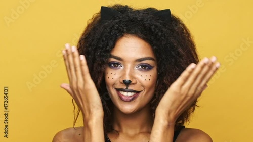 Young african lady with cat make-up and wearing black dress hiding her face and showing different emotions idolated over yellow photo