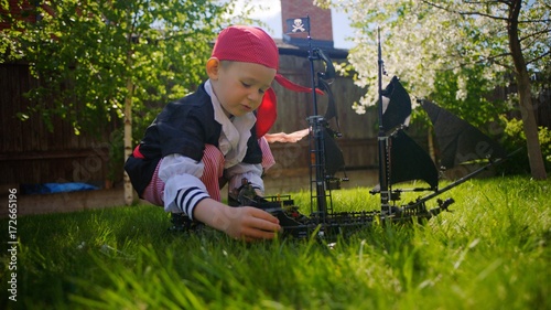 Little boy wearing pirate costume is playing in toy ship of pirates of caribbean on the lawn on Halloween