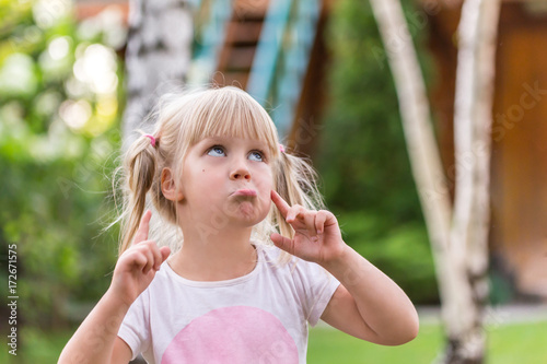 Portrait of cute adorable little blonde caucasian girl in a white T-shirt in a park, looking up, thinking or dreaming of something, puzzled or astonished, happy childhood concept.