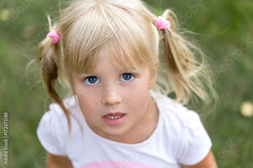 Close up portrait of cute adorable little blonde caucasian girl in a white T-shirt in a park, looking aside, thinking or dreaming of something, puzzled or astonished, happy childhood concept.