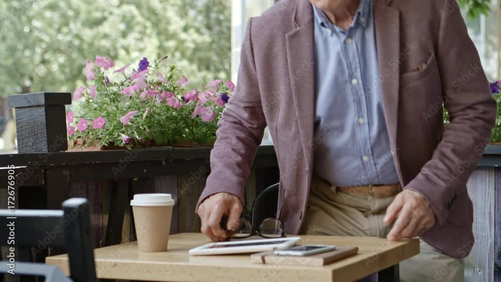 Senior businessman walking and sitting at table in outdoor restaurant, putting on eyeglasses and turning on digital tablet Senior businessman walking and sitting at table in outdoor restaurant, putting on eyeglasses and turning on digital tablet