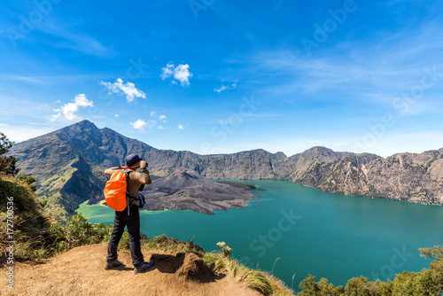Hiker man with orange backpack standing, enjoy and happy with active volcano Baru Jari, Lake Segara Anak and summit of Mount Rinjani view after finished climbing at Rinjani mountain, Lombok, Indonesia