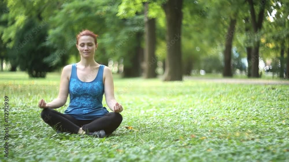 Full of joy young women wearing sports clothes blue shirt and black tights in lotus position meditating in the quiet scenery of late summer in the park, camera is gently moving / Joy of doing yoga Full of joy young women wearing sports clothes blue shirt and black tights in lotus position meditating in the quiet scenery of late summer in the park, camera is gently moving / Joy of doing yoga