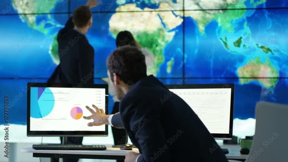 Business team discussing large world map graphic on video wall, with pie charts & graphs displayed on computer screen. Global business concept. Business team discussing large world map graphic on video wall, with pie charts & graphs displayed on computer screen. Global business concept.