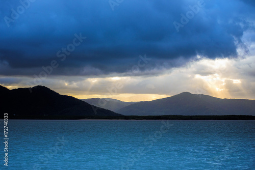 Quiet sea with mountains in the background and crepuscular sun rays shining through the clouds. Landscape of Australia