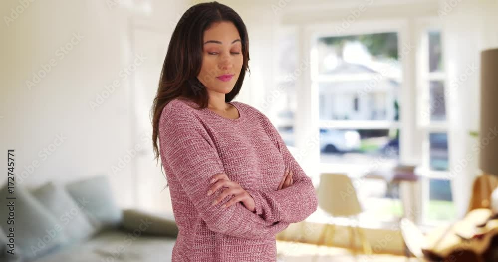 Portrait of young Latina woman in cozy pink sweater smiling at camera with arms crossed. Attractive female dressed casually looking at camera with friendly expression inside bright living room. 4k 