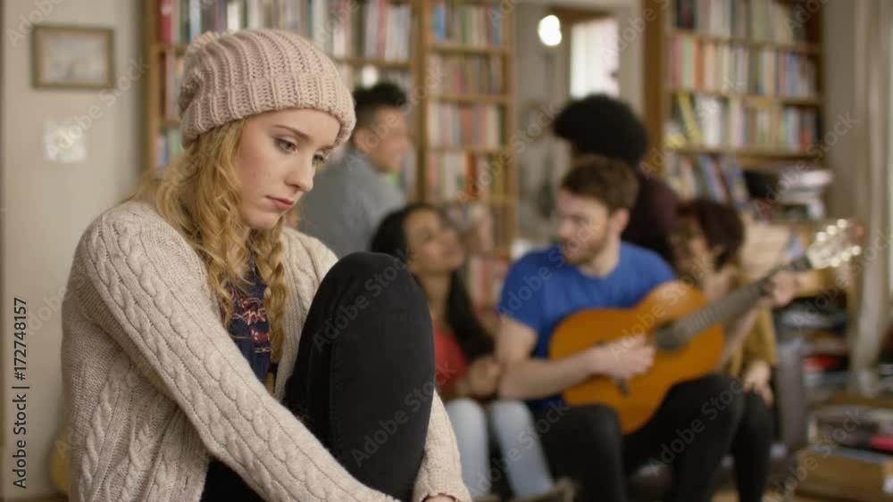 Young woman in shared apartment feeling sad and isolated while others socialize in background. Young woman in shared apartment feeling sad and isolated while others socialize in background.