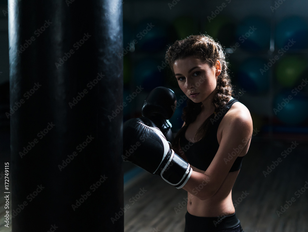 Young fighter boxer girl wearing boxing gloves in training with heavy punching bag.