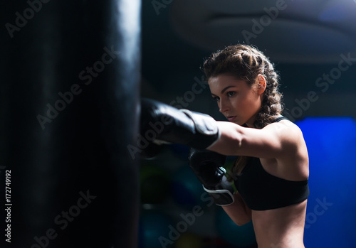 Young fighter boxer fit girl wearing boxing gloves in training with heavy punching bag.