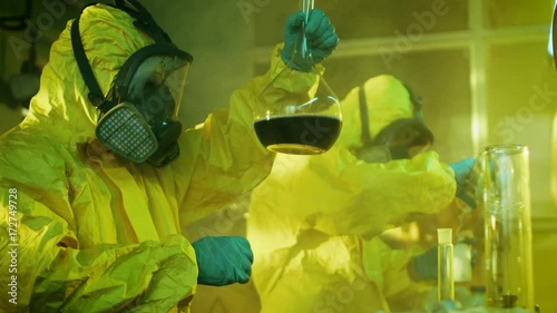 In the Underground Drug Laboratory Team of Clandestine Chemists Synthesises Illegal Drugs, One Holds Beaker with Chemicals and Checks it's Consistency. They Cook Drugs in the Abandoned Building. photo