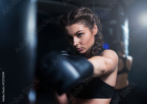 Young fighter boxer fit girl wearing boxing gloves in training with heavy punching bag in gym. Low key image. Woman power. Moment of punch