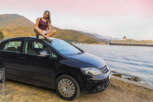 девушка сидит на крыше автомобиля на фоне моря в Европе 