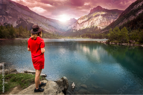 Une jeune femme blonde de dos avec une robe rouge et un chapeau au bord d'un lac de montagne sous un coucher de soleil