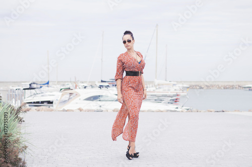 Young fashionable brunette woman wearing long dress in sunglasses posing near sea, pier with yachts